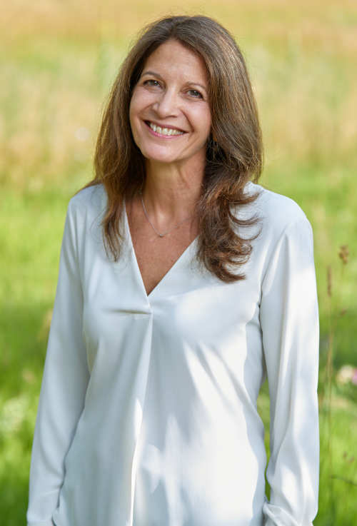 Photo of Michelle Decola, wearing a white blouse, standing outside in a green field, smiling.