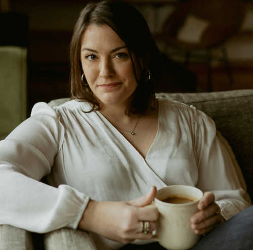 Photo of Therapist, Katie Schuh, sitting with a cup of coffee, smiling, wearing a white sweater.