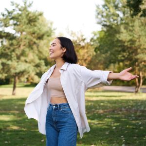 Westside Behavioral Care ADHD and ADD Section - Young woman in jeans and a white shirt smiling with her arms outstretched in a green park surrounded by trees on a sunny day.