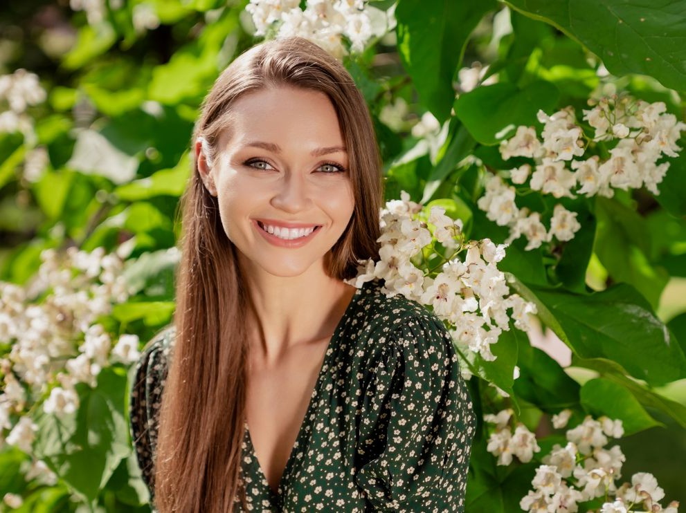Westside Behavioral Care About Westside Section - Young woman with long brown hair smiling in front of lush green leaves and white blooming flowers on a sunny day.