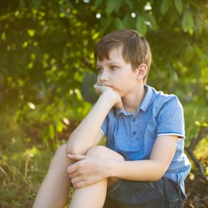 Westside Behavioral Care Anxiety Section - Young boy in a blue shirt sitting on the ground in a shaded wooded area, resting his chin on his hand with a thoughtful expression.