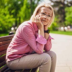Westside Behavioral Care Depression Section - Thoughtful older woman in glasses and a pink shirt sitting on a park bench, resting her face in her hands with a gentle expression.