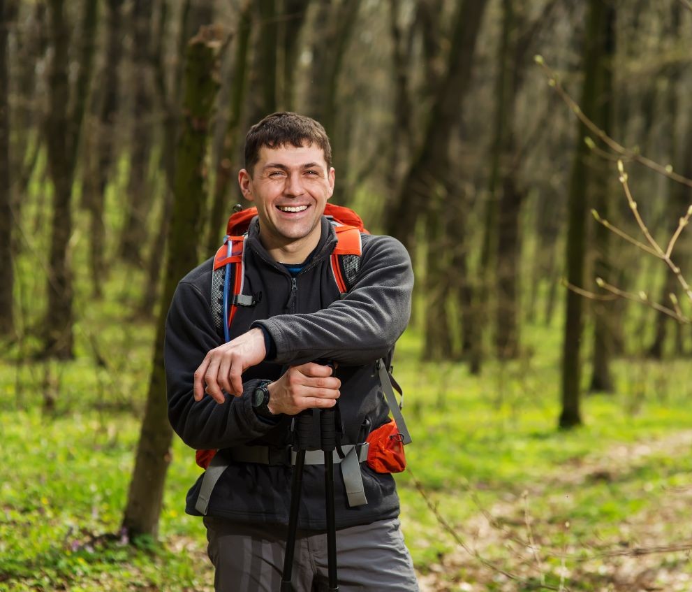 Westside Behavioral Care Meet our Kaiser Prescribers Section - Happy man in hiking gear with a red backpack and trekking poles standing on a trail in a forest with fresh green foliage.