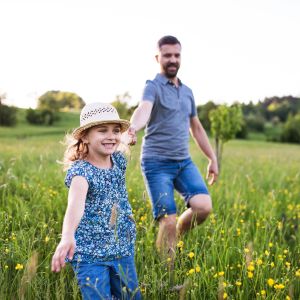 Westside Behavioral Care Parenting Skills Section - Young girl wearing a sunhat and floral top running joyfully through a wildflower field while holding hands with her smiling father behind her.